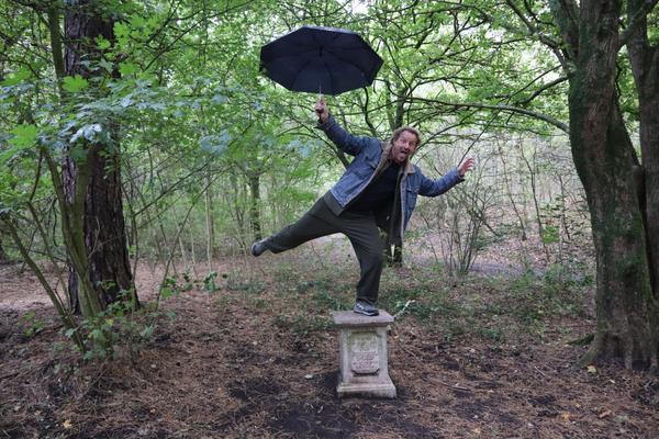 A man playfully balances on a stone pedestal in a lush green forest, holding an open black umbrella above his head